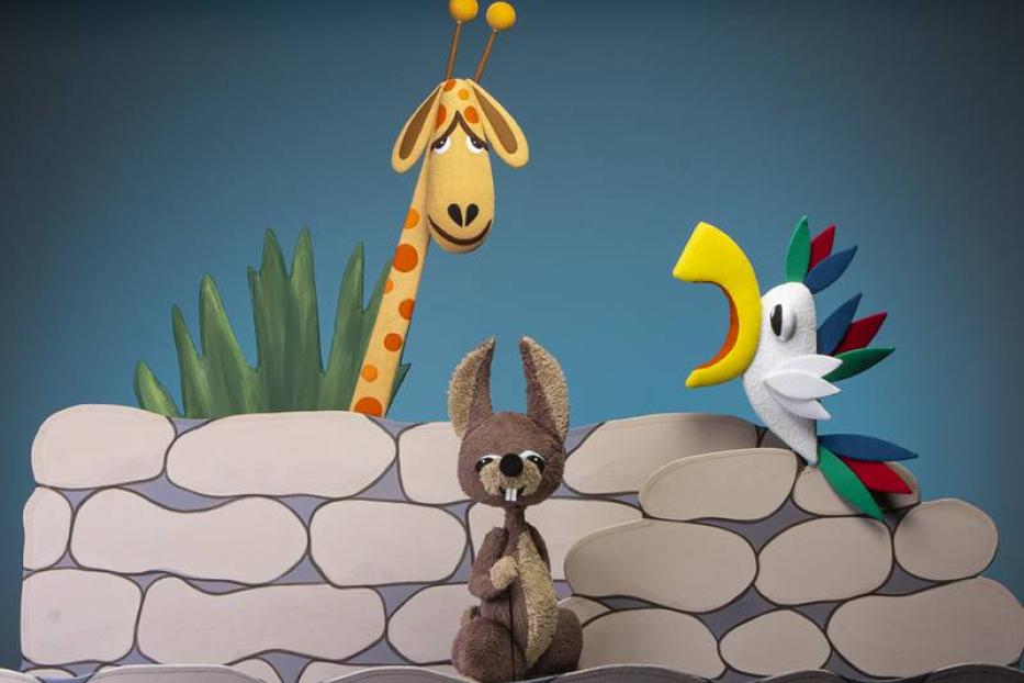 Eine Giraffe, ein Hase und ein Papagei sitzen hinter einer Steinmauer.