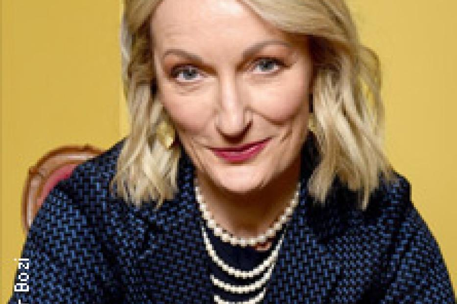 Eine blonde Frau mit Perlenkette lächelt in die Kamera.