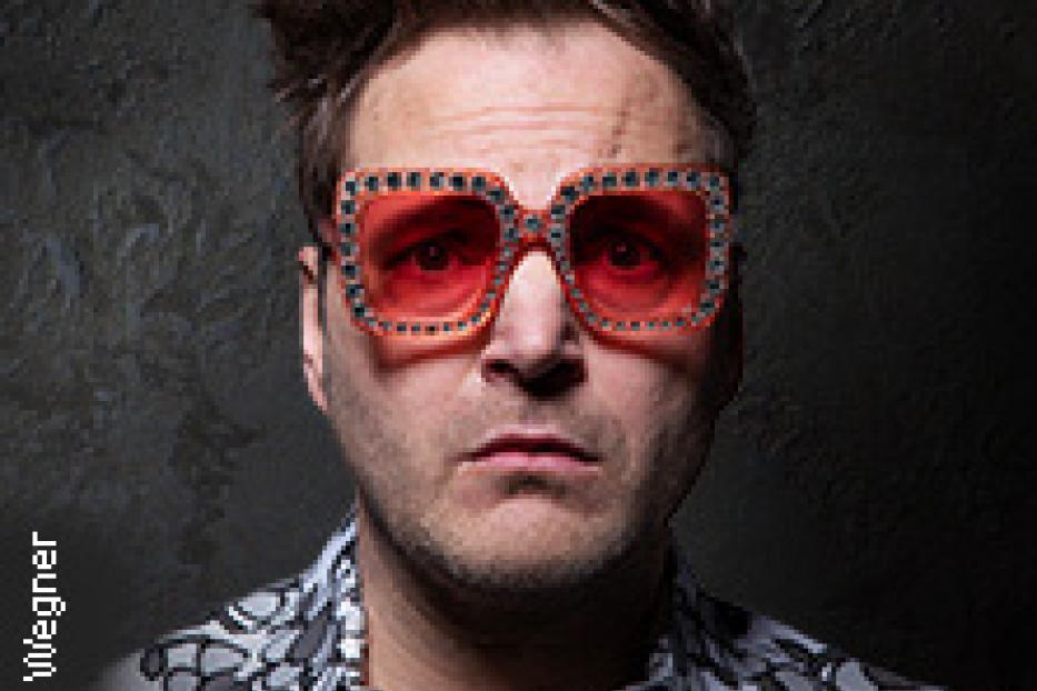 Ein Mann mit auffälliger Brille und gemustertem Hemd blickt in die Kamera.