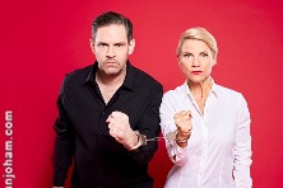 Ein Mann und eine Frau stehen vor einem roten Hintergrund, ihre Hände sind mit Handschellen gefesselt.