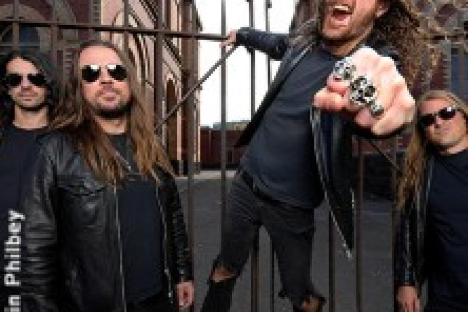 Die Band Airbourne posiert vor einem Tor mit Backsteingebäuden im Hintergrund.