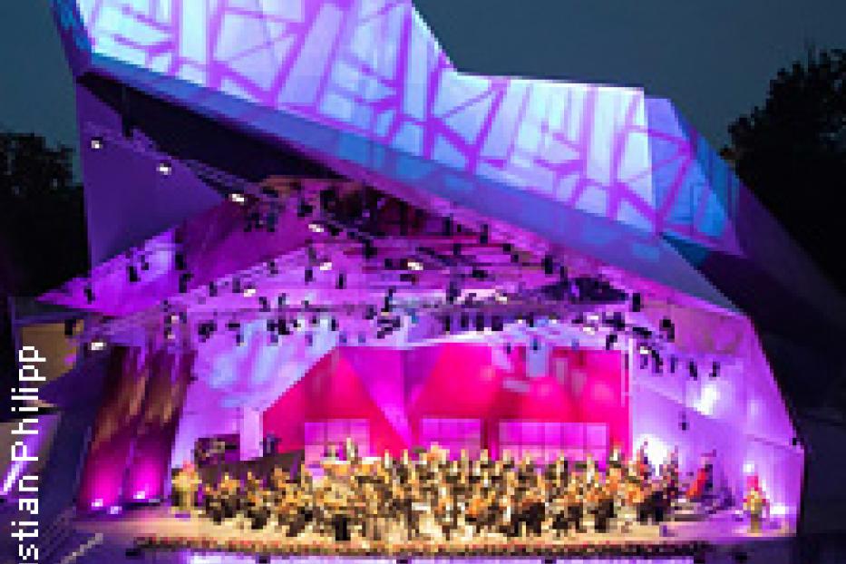 Ein Orchester spielt auf einer beleuchteten Bühne vor einem Publikum.