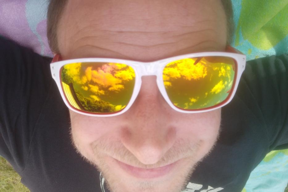 Ein Mann mit Sonnenbrille liegt im Freien, der Himmel spiegelt sich in den Gläsern.