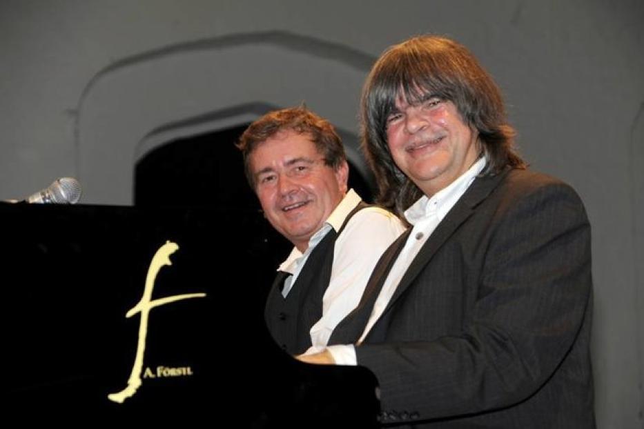 Zwei Männer stehen lächelnd an einem Klavier.