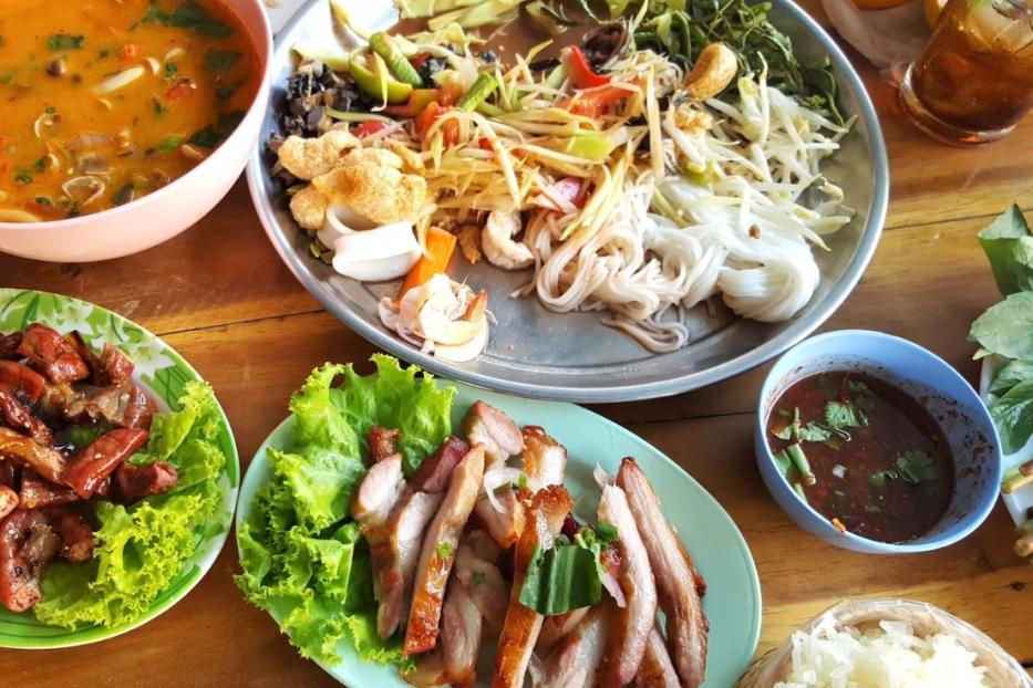 Verschiedene thailändische Gerichte, darunter Suppe, Salat und gegrilltes Fleisch, werden auf einem Tisch präsentiert.