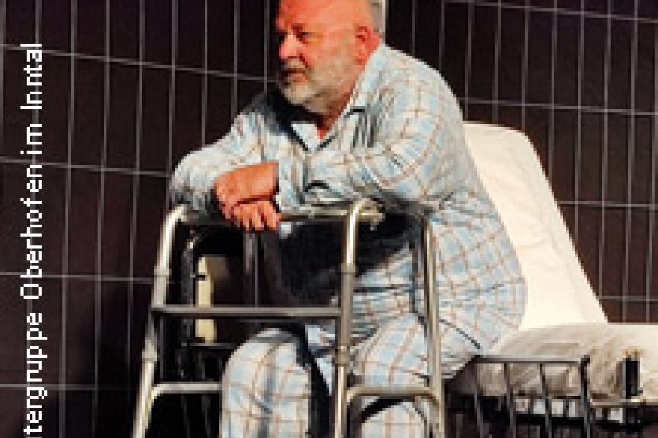 Ein älterer Mann im Pyjama sitzt mit einem Rollator vor einem Krankenbett.