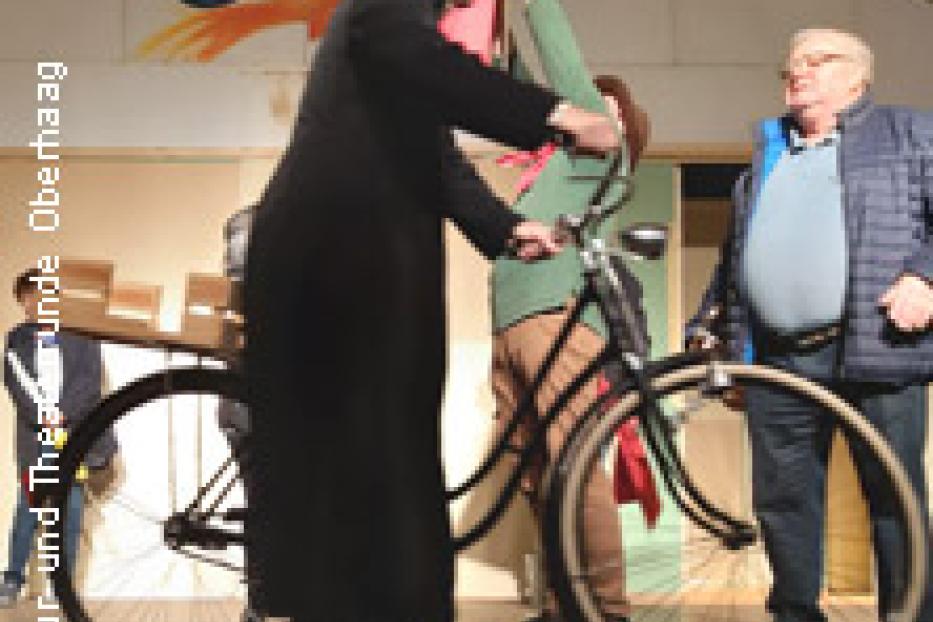Ein als Priester verkleideter Mann steht neben einem Fahrrad auf einer Bühne.