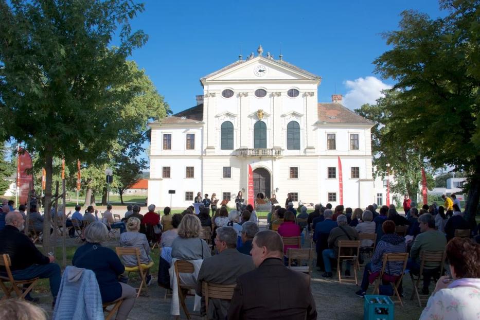 Zuschauer sitzen vor einem weißen Gebäude und hören einer Musikgruppe zu.