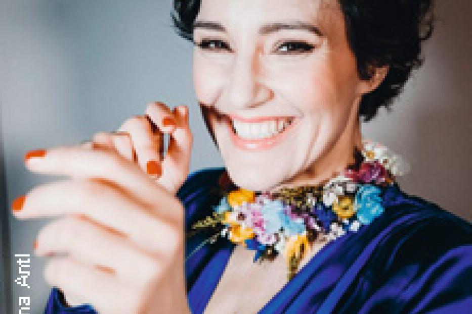 Eine lächelnde Frau mit dunklen, kurzen Haaren und einem blauen Kleid mit Blumenkragen.