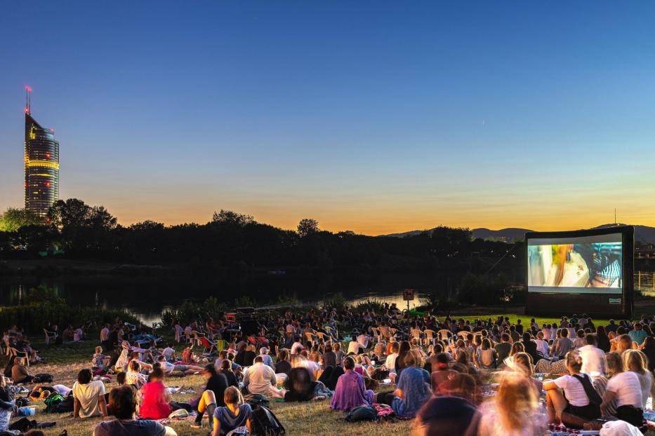 Eine Menschenmenge genießt ein Open-Air-Kino mit Blick auf den Donauturm bei Sonnenuntergang.