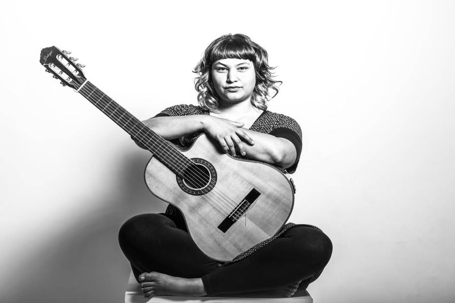 Eine Frau sitzt mit einer Gitarre im Schneidersitz auf einem Stuhl.