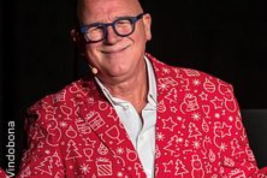 Ein Mann mit Brille und rotem Sakko mit Weihnachtsmotiven lächelt.
