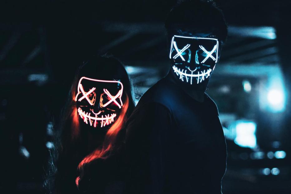 Zwei Personen tragen leuchtende Masken mit einem „X“ für die Augen und einem grinsenden Mund.