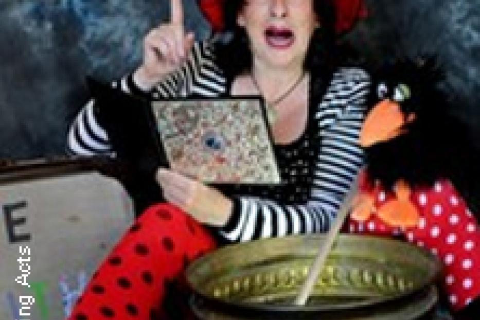 Eine Frau mit einem roten Hut und gepunkteten Leggings sitzt neben einem Kessel und einer Puppe.