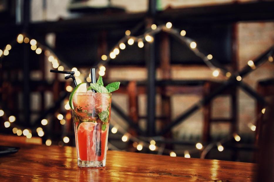 Ein leicht rötlicher Drink mit Limetten, Minze, Eiswürfel und Strohhalm, allein stehend auf einem Tisch, dahinter ein Hintergrund mit Lichterketten