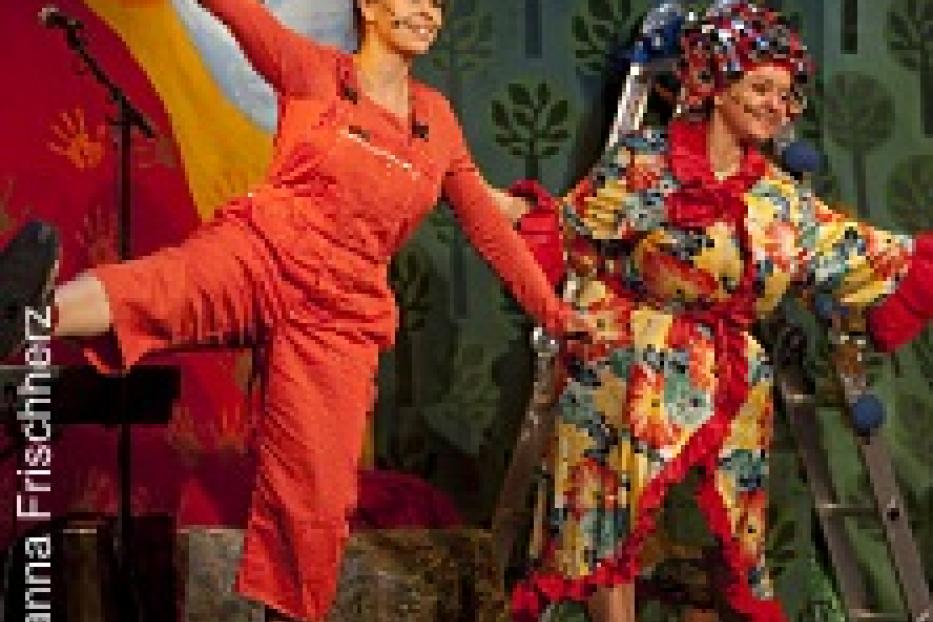 Zwei Schauspielerinnen in bunten Kostümen stehen auf einer Bühne vor einer gemalten Kulisse.