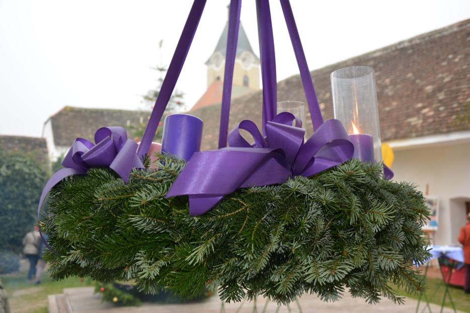 Ein hängender Adventskranz mit lila Kerzen und Schleifen vor einem Gebäude.