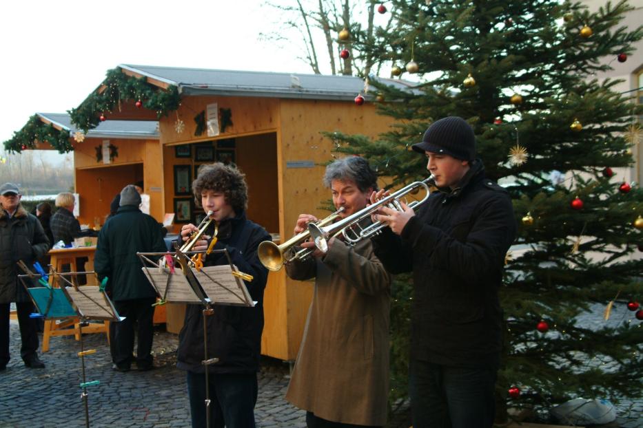 Drei Personen spielen Trompete vor einem Weihnachtsbaum auf einem Weihnachtsmarkt.