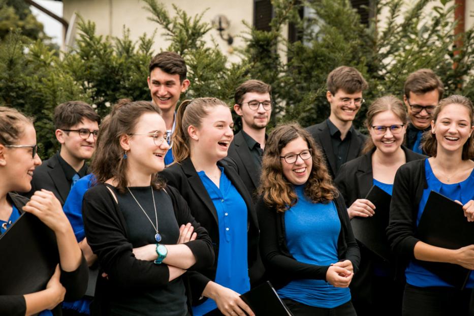 Eine Gruppe junger Leute, vermutlich ein Chor, steht lachend vor einem grünen Hintergrund.