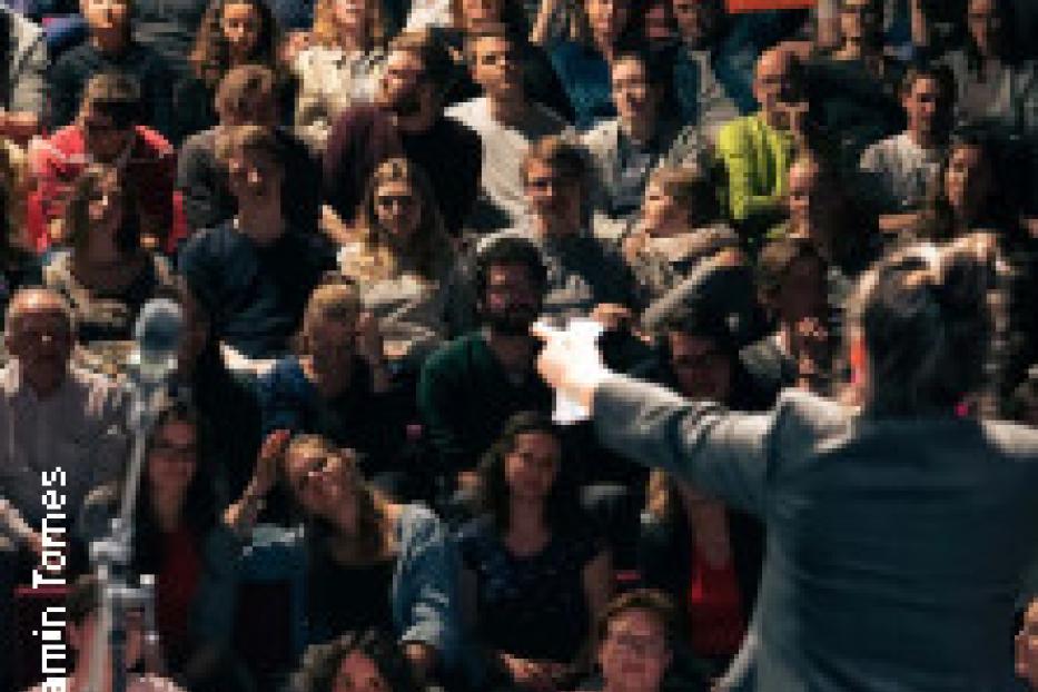 Eine Menschenmenge sitzt in einem dunklen Raum und schaut auf eine Person, die vor ihnen steht.