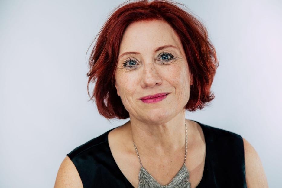 Eine Frau mit roten Haaren und einem schwarzen Oberteil lächelt in die Kamera.