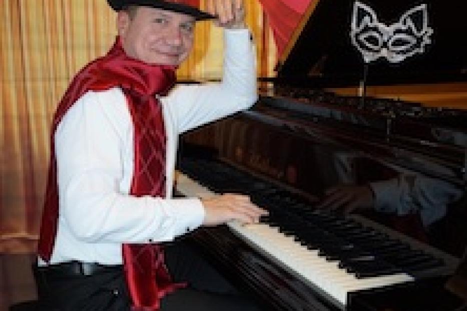 Ein Mann mit rotem Zylinder und Schal spielt Klavier; eine Maske steht auf dem Klavier.