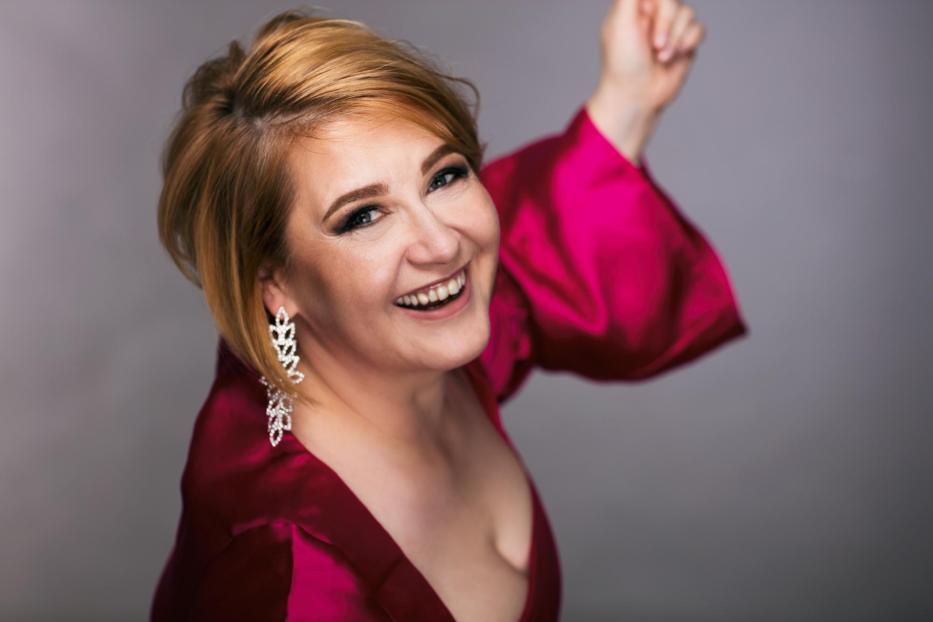 Eine lachende Frau mit Ohrringen und einem pinkfarbenen Kleid posiert vor einem grauen Hintergrund.