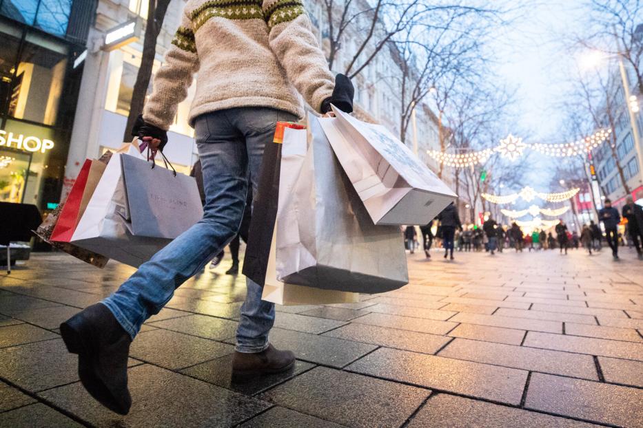 Eine Person trägt mehrere Einkaufstaschen auf einer belebten, festlich beleuchteten Einkaufsstraße.