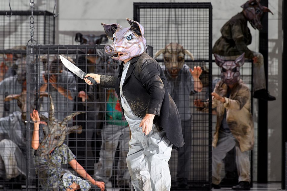 Ein Mann mit Schweinsmaske hält ein Messer, während andere Personen mit Tiermasken in Käfigen gefangen sind.