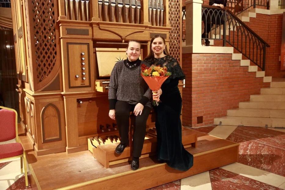 Zwei Frauen posieren vor einer Orgel, eine hält einen Blumenstrauß.
