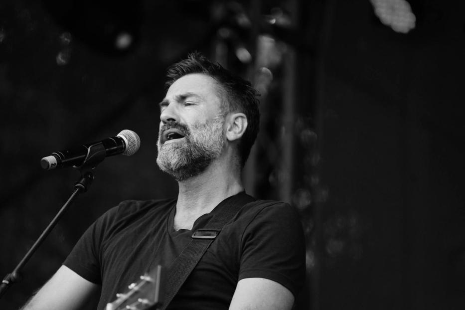 Ein bärtiger Mann singt mit Gitarre in ein Mikrofon.