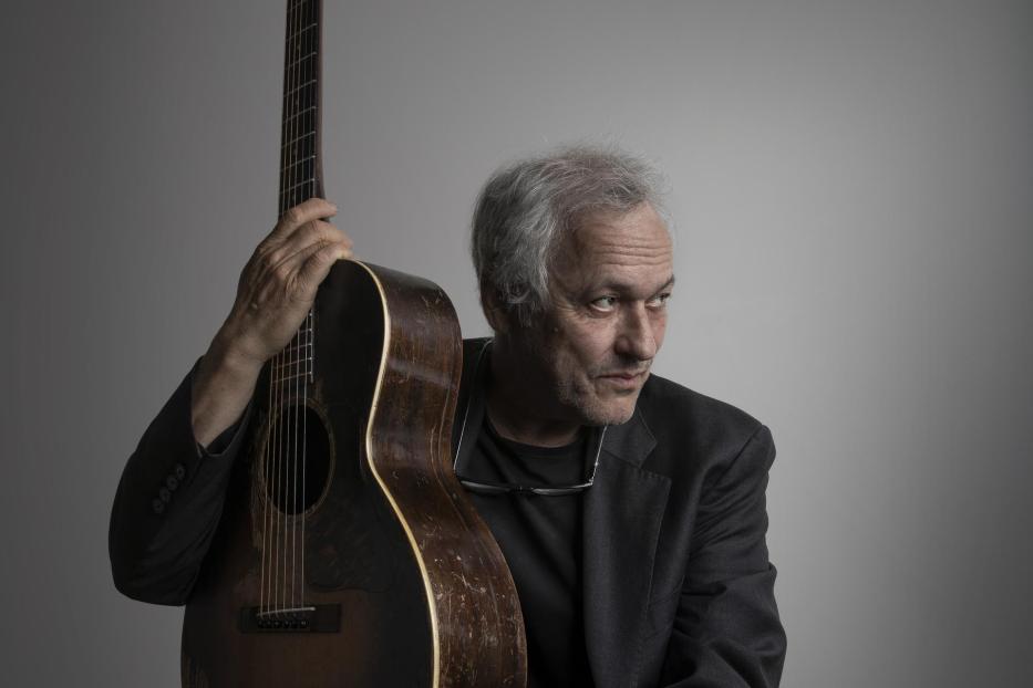 Ein Mann mit grauen Haaren hält eine akustische Gitarre.