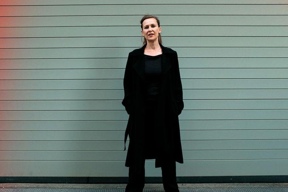 Eine Frau in einem schwarzen Mantel steht vor einer grauen Wand.
