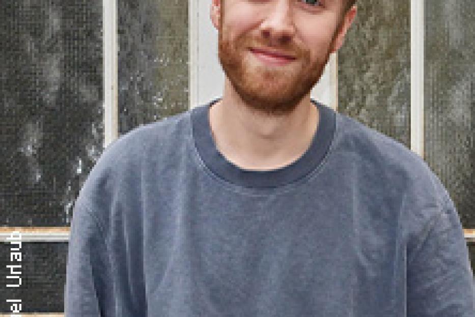 Ein junger Mann mit Bart und blauem Pullover lächelt in die Kamera.
