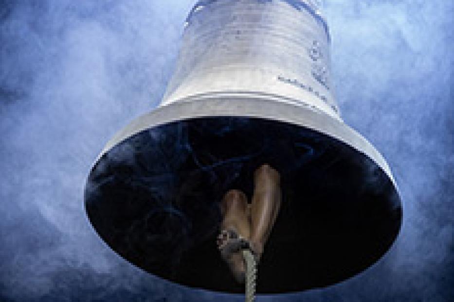 Eine große Glocke hängt an einem Seil vor einem dunklen, nebligen Hintergrund.