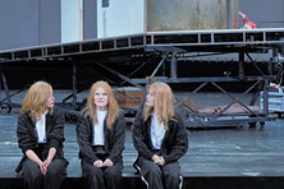 Drei junge Frauen in schwarzen Anzügen sitzen auf einer Bühne vor einer industriellen Kulisse.