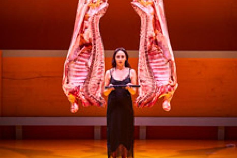 Eine Frau in einem schwarzen Kleid steht zwischen zwei aufgehängten Tierkadavern.