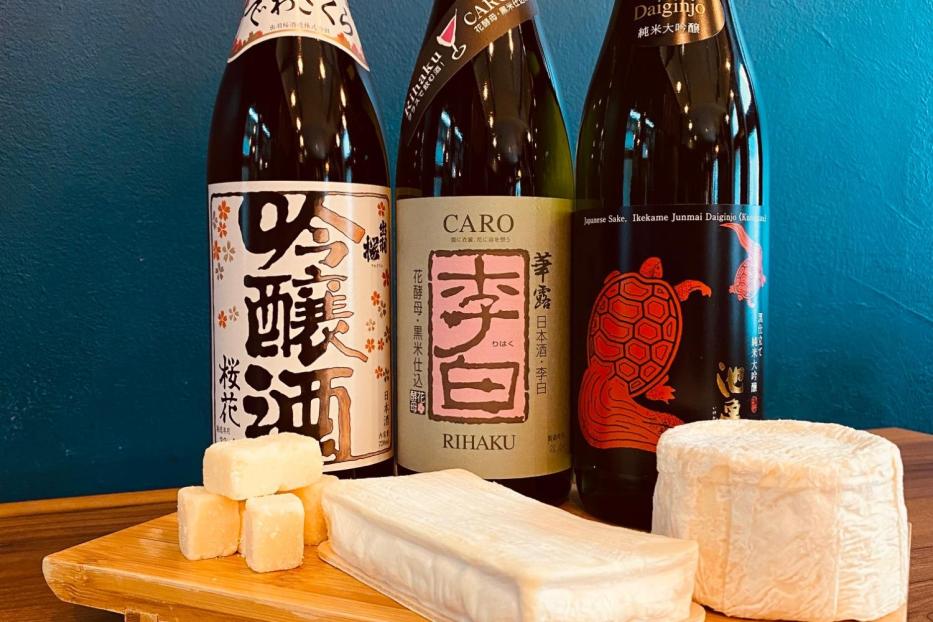 Drei Flaschen japanischer Sake und verschiedene Käsesorten auf einem Holzbrett.