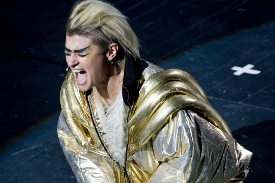 Eine Person in goldener Kleidung kniet auf der Bühne und schreit.