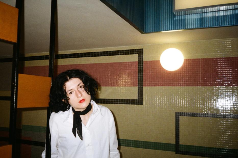 Eine junge Frau mit dunklen Haaren und rotem Lippenstift steht vor einer Wand mit bunten Fliesen.