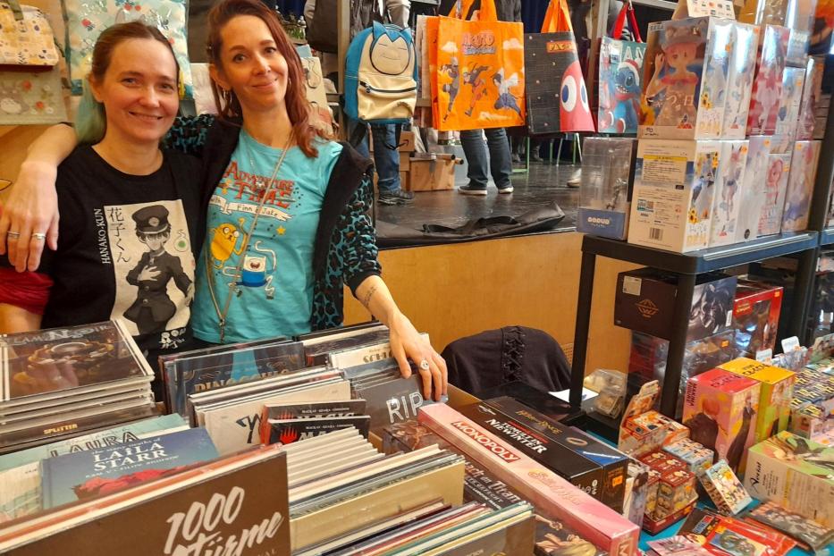 Zwei Frauen posieren an einem Stand mit Comics, Filmen und Merchandise auf einer Comic- und Filmbörse.