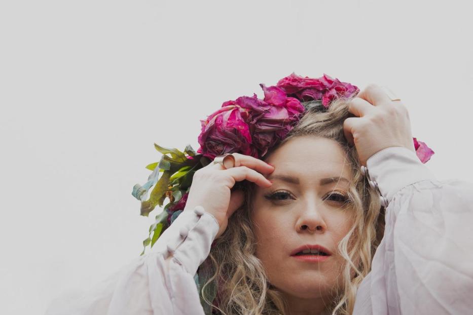 Eine Frau mit blonden Locken und einem Blumenkranz blickt in die Kamera.