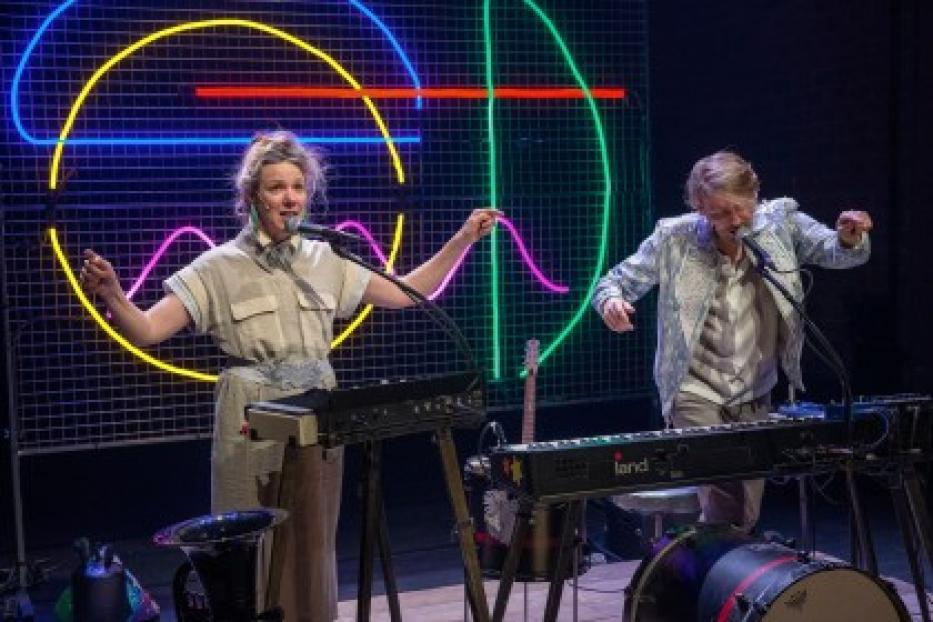 Zwei Musiker treten mit Keyboards und Mikrofonen vor einem neonbeleuchteten Hintergrund auf.
