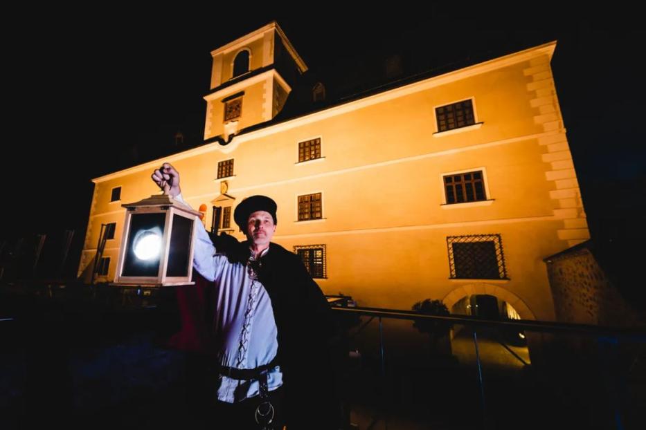 Ein Mann in historischer Kleidung hält eine Laterne vor einem gelb beleuchteten Gebäude.