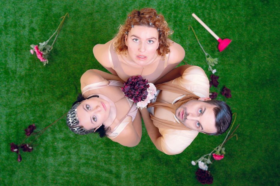 Drei Personen liegen auf einer grünen Oberfläche, umgeben von Blumen und einem Pömpel.