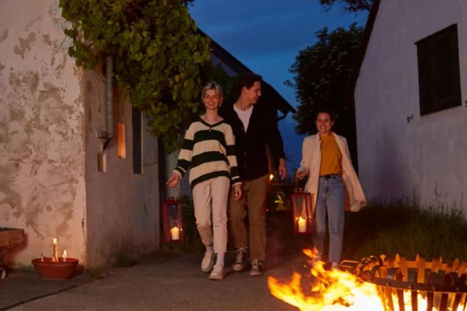 Drei junge Leute spazieren mit Laternen an einem Lagerfeuer vorbei.