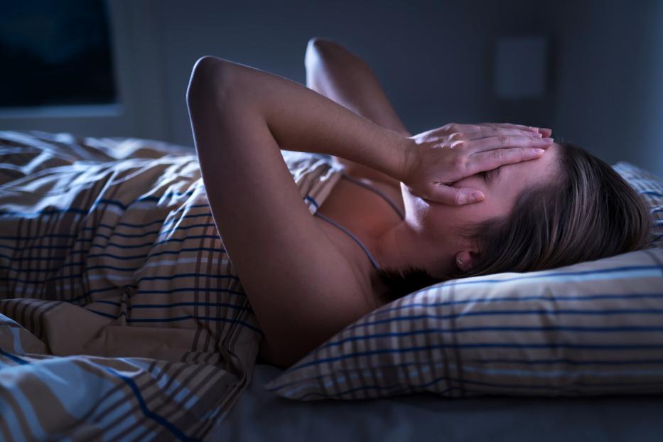 Eine Frau liegt im Bett und bedeckt ihr Gesicht mit den Händen, wirkt erschöpft oder gestresst.