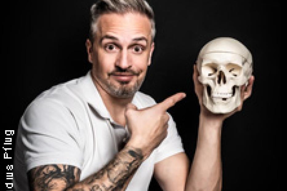 Ein Mann mit Tätowierungen zeigt auf einen menschlichen Schädel.