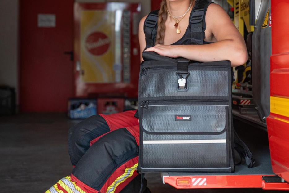 Eine lächelnde Feuerwehrfrau sitzt mit einer Tasche auf dem Trittbrett eines Feuerwehrautos.