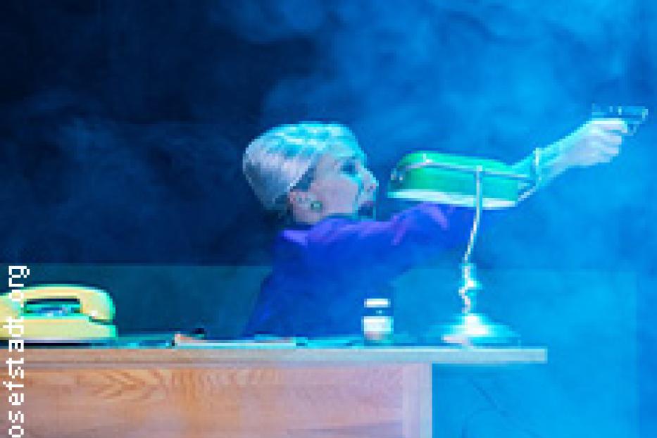 Eine Frau mit weißem Haar zielt mit einer Pistole über einen Schreibtisch.